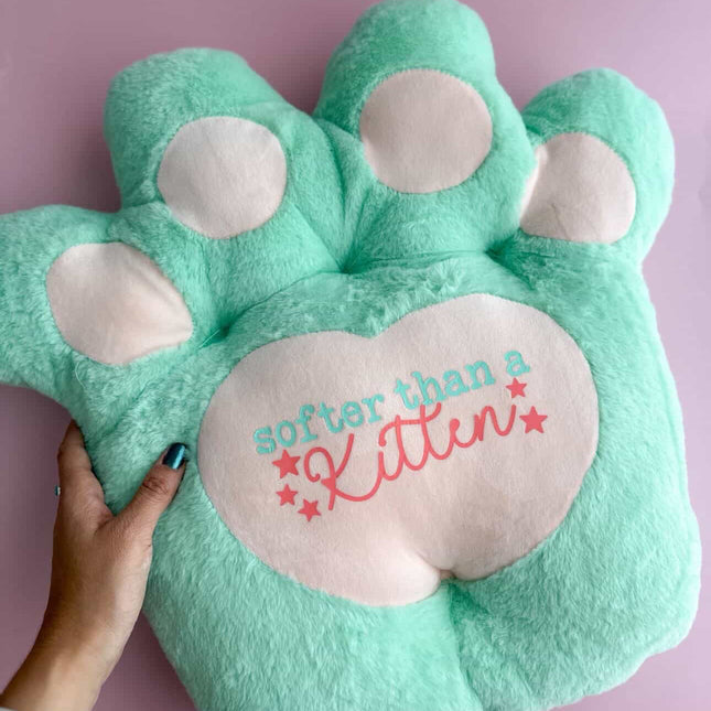 Green paw-shaped pillow with 'softer than a kitten' text on a pink background